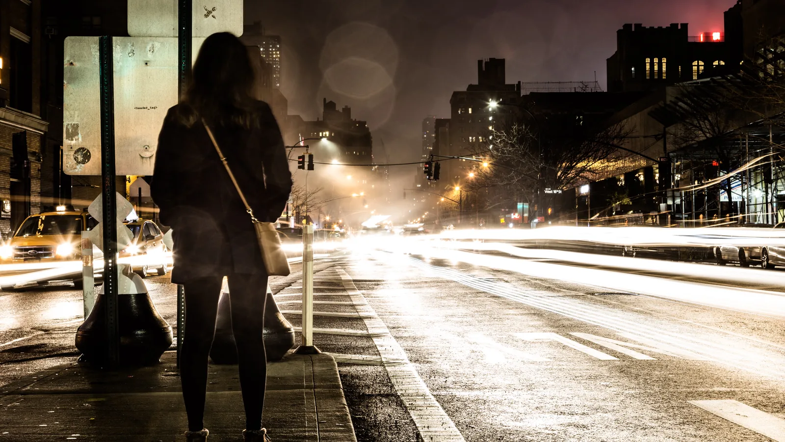 Une passante seule au bord d'une avenue la nuit, les phares filent derrière elle