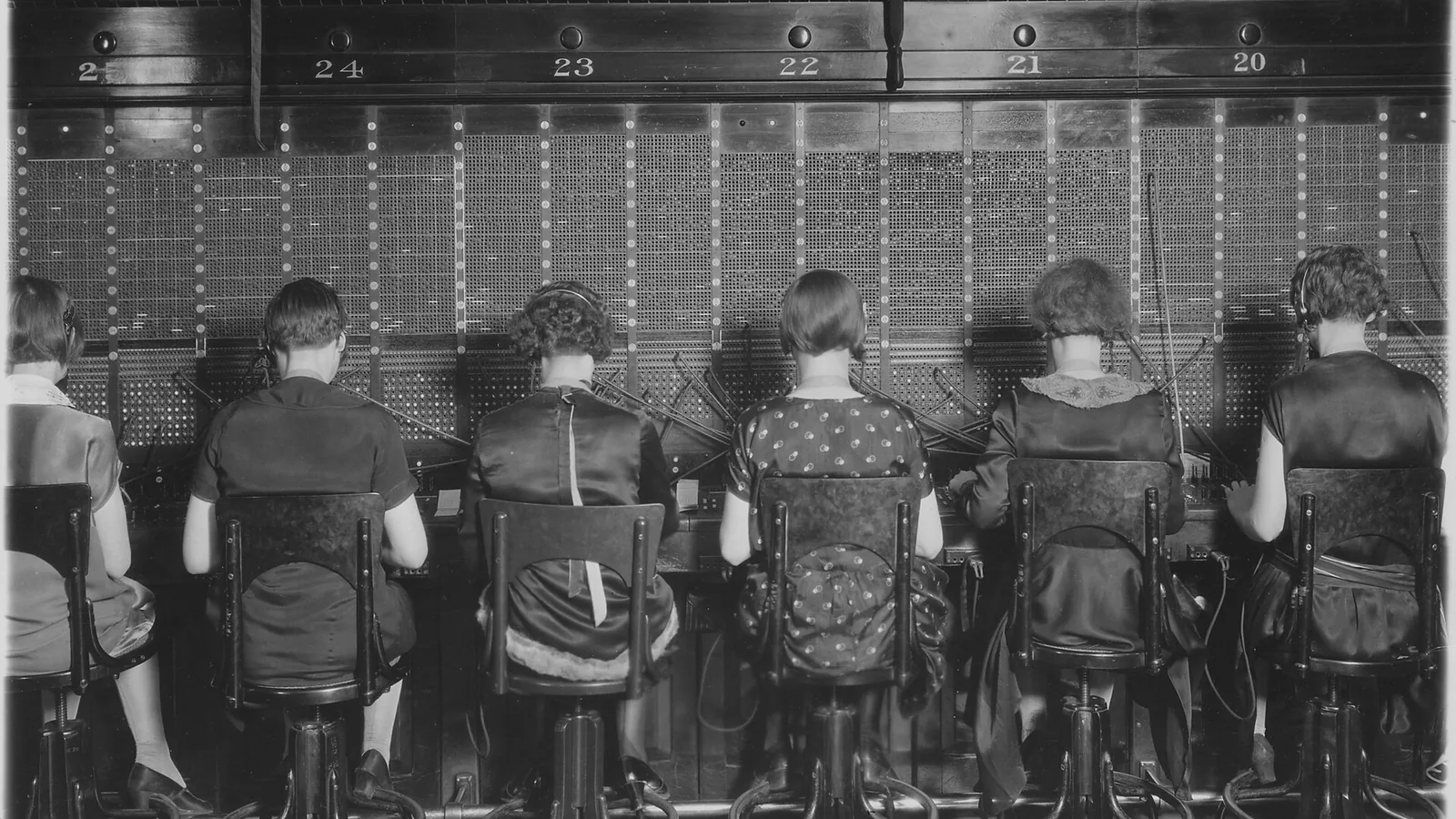 Une rangée de standardistes de dos devant un tableau de connexions téléphoniques